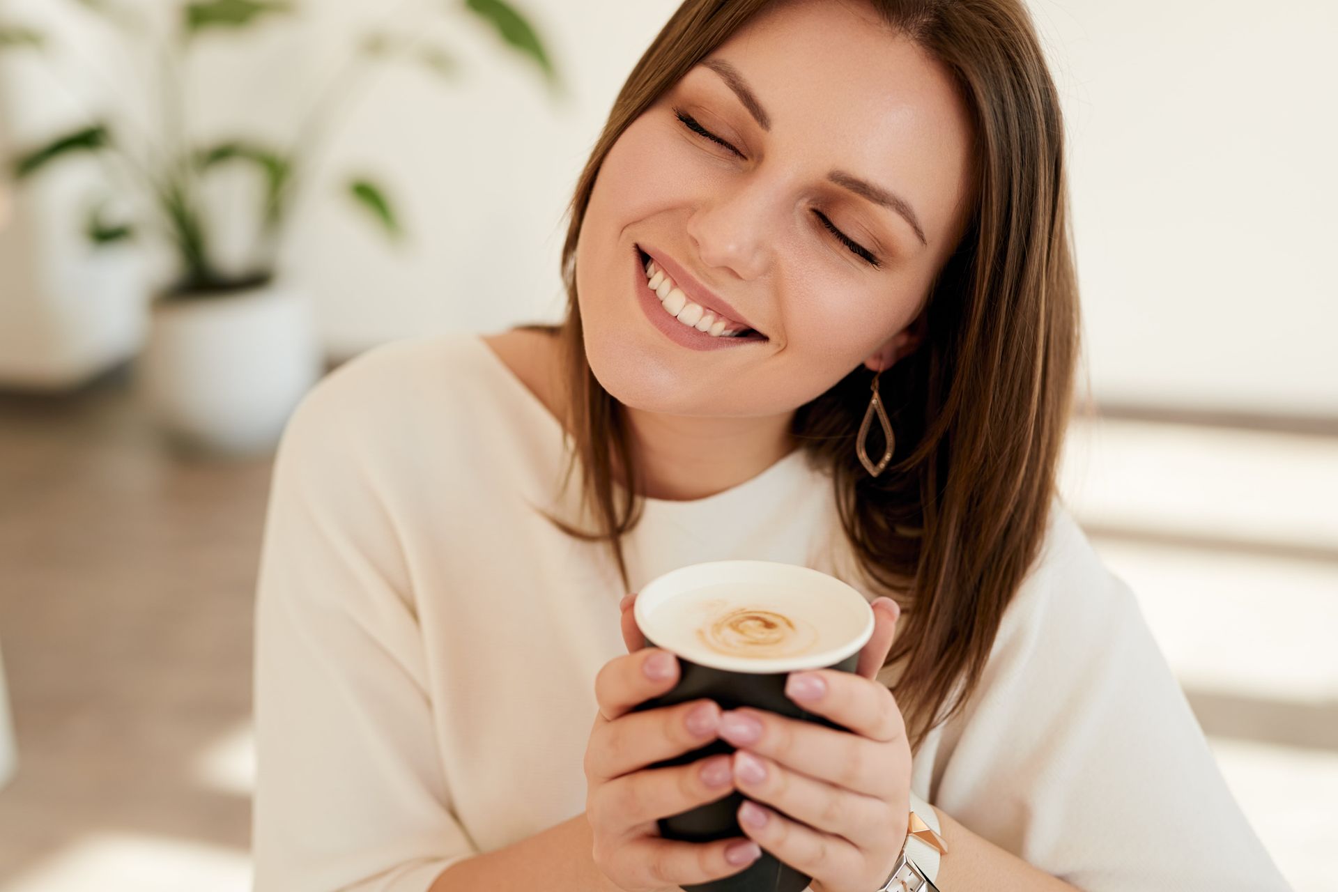 Woman smiles with eyes closed, holding a mug of coffee in a bright room.