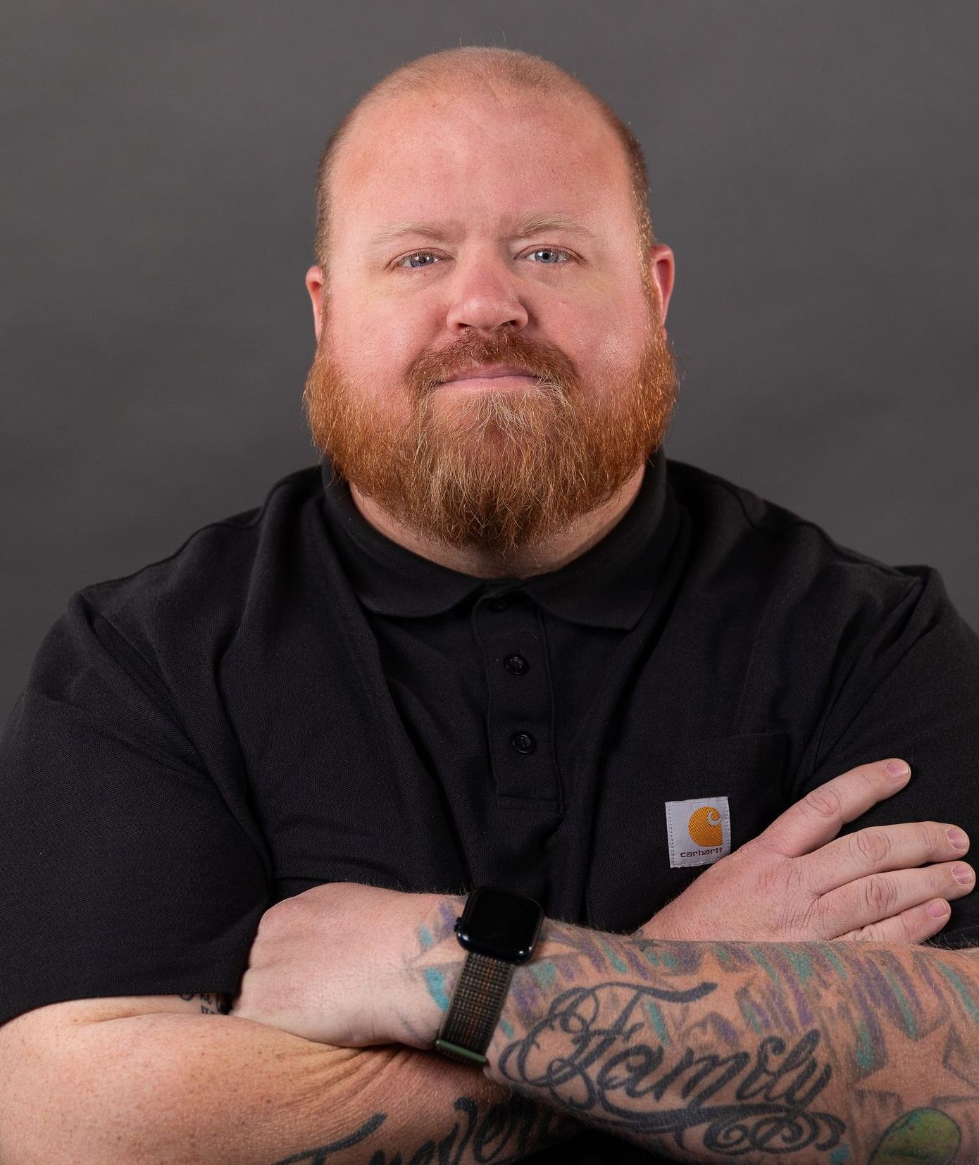 A man with a beard and tattoos on his arms is standing with his arms crossed.