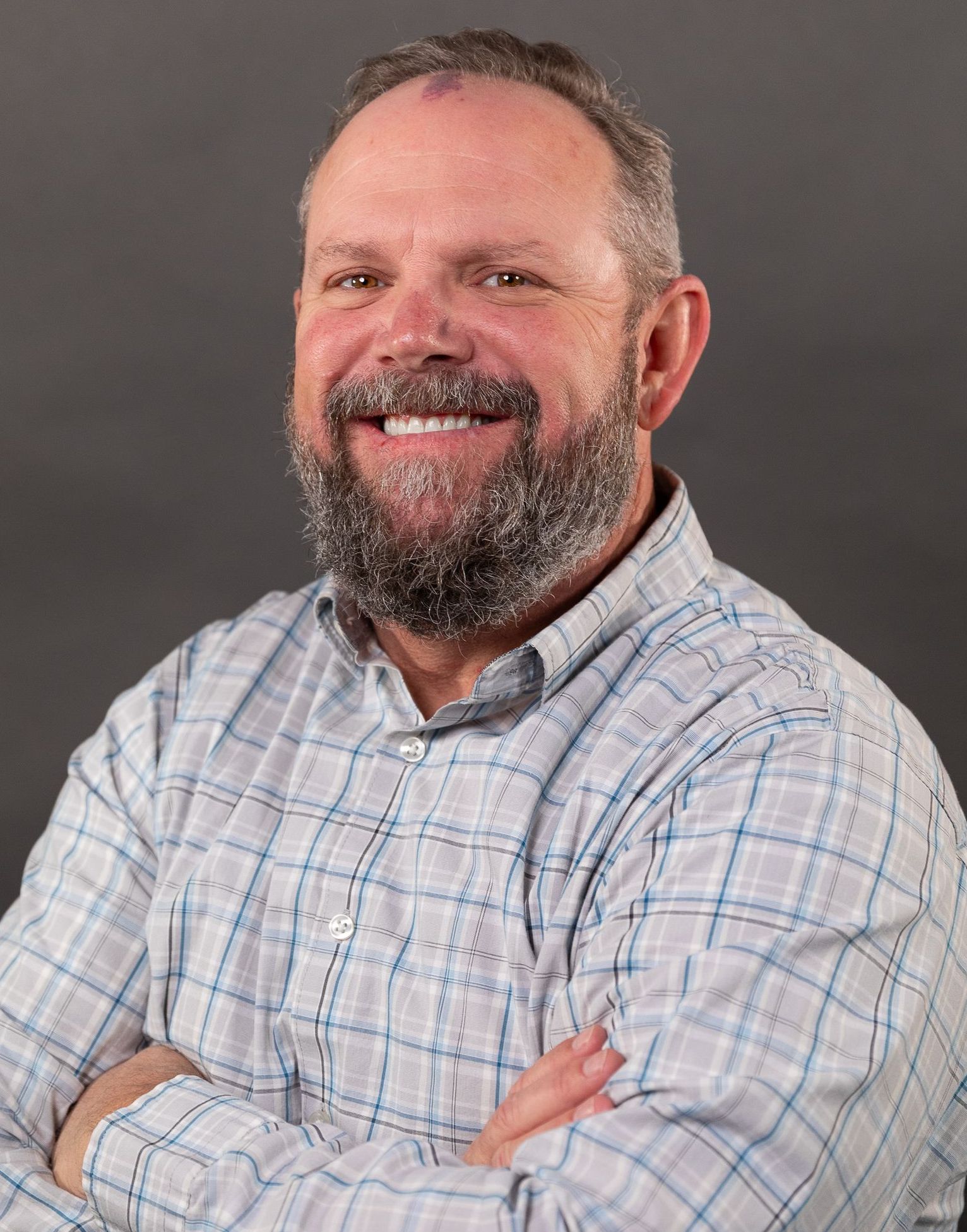A man with a beard and a plaid shirt is smiling with his arms crossed.