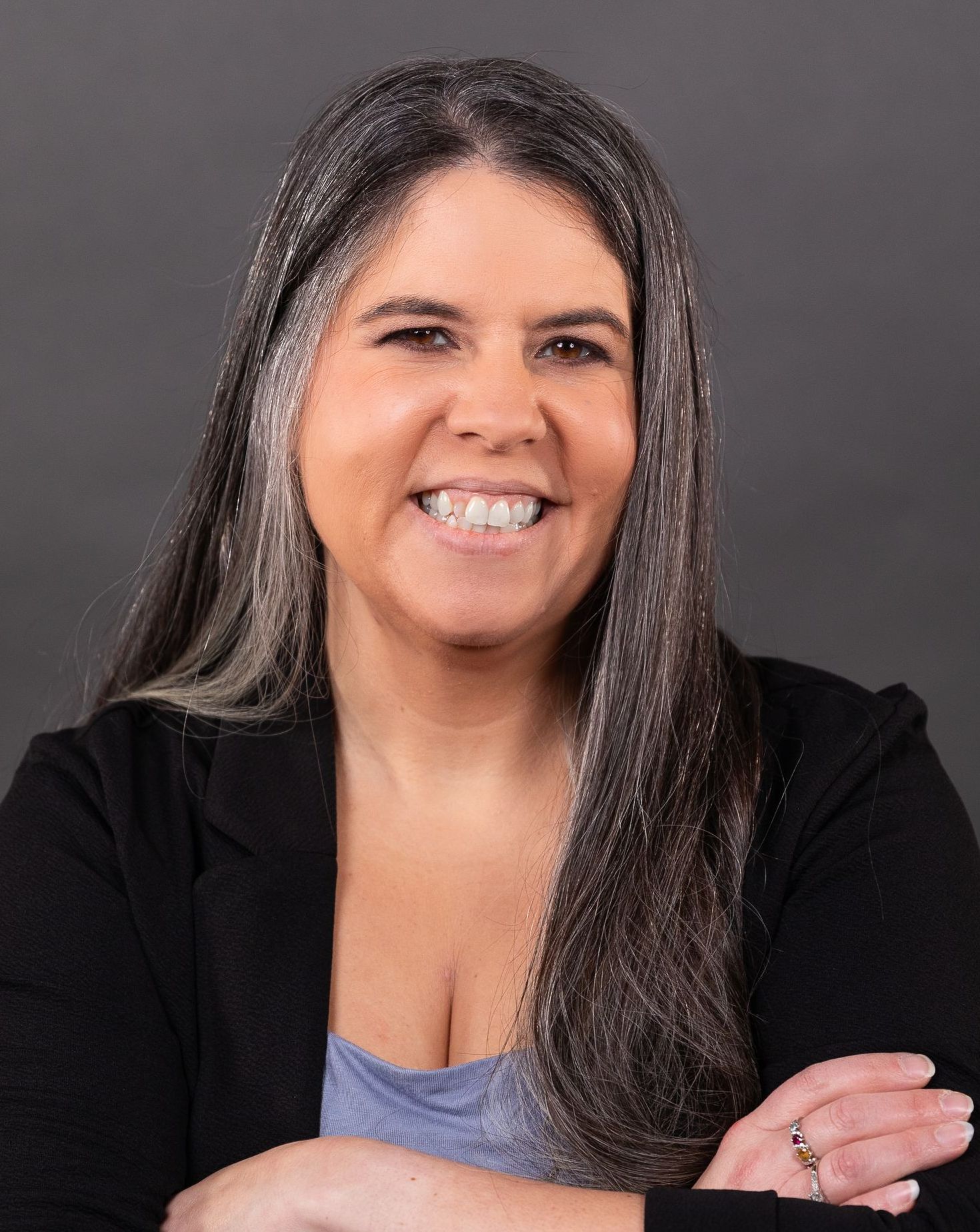 A woman with long gray hair is smiling with her arms crossed.