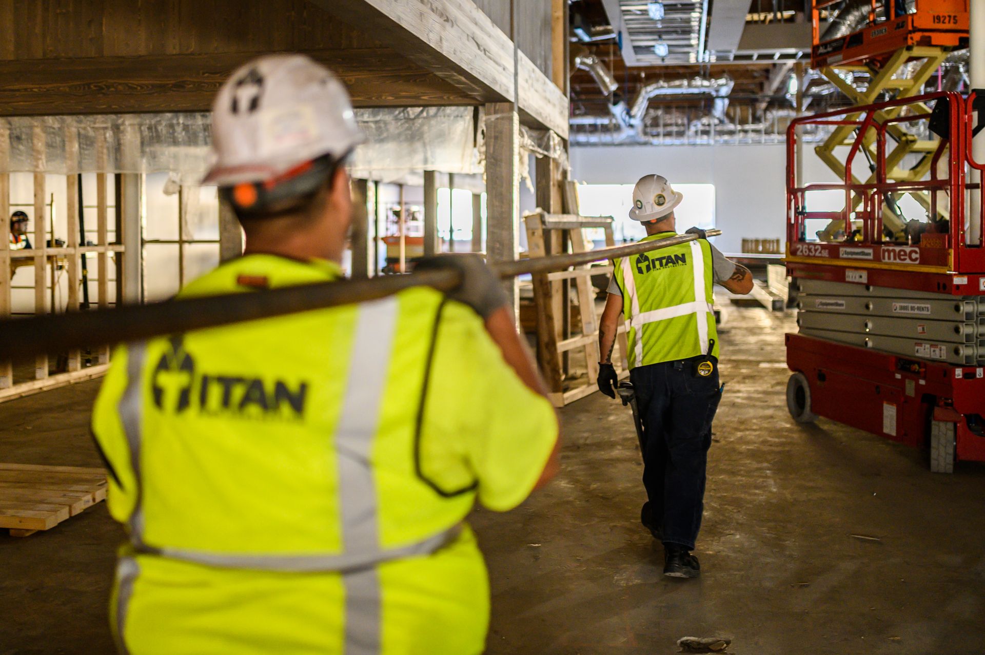Two construction workers in neon vests carrying lumber in a building. One worker's back is to the camera.