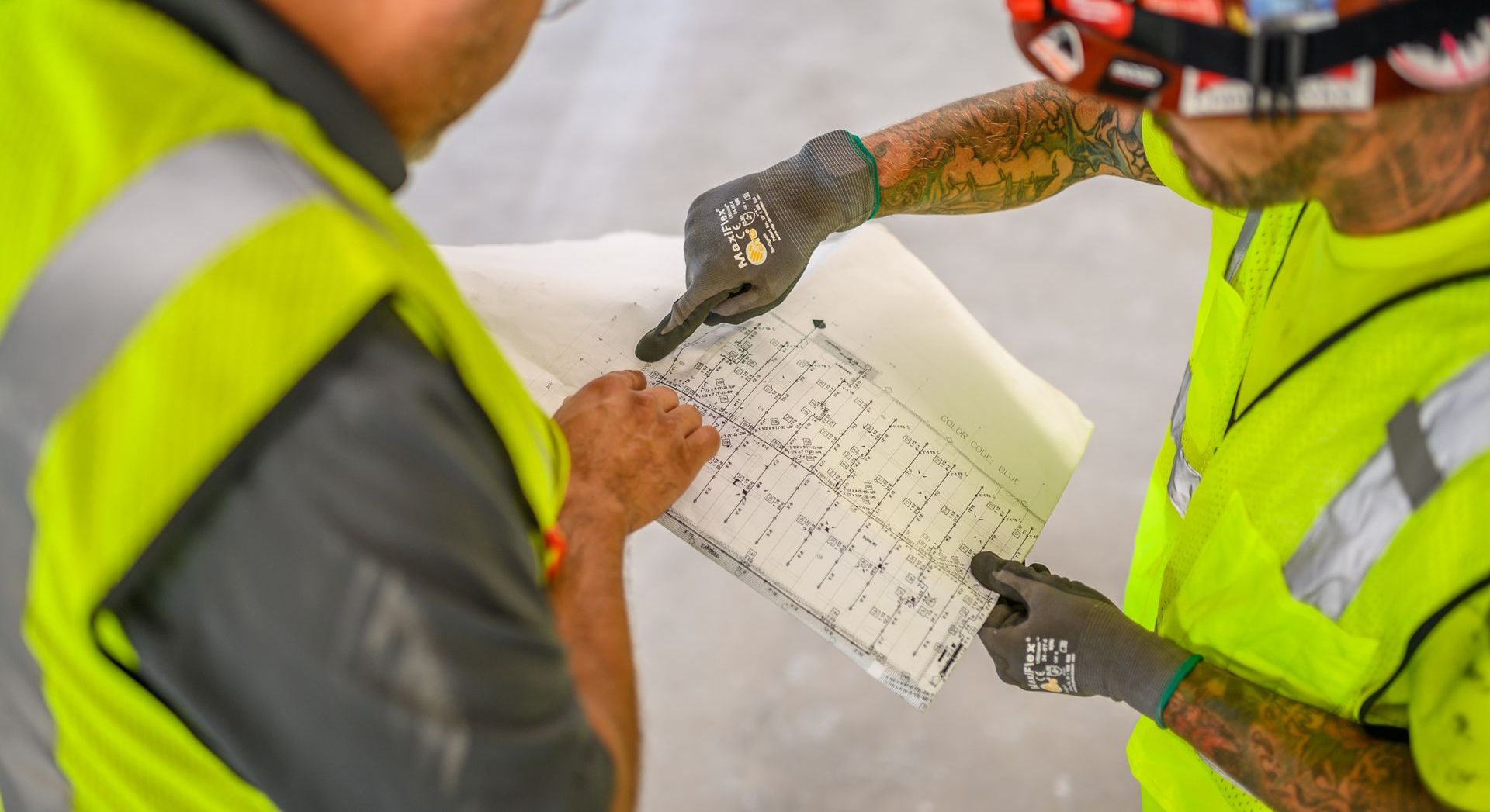 Two construction workers examining blueprints, wearing safety vests and gloves.