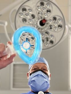 Person in surgical attire being prepped with an anesthesia mask under an operating room light.