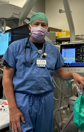 A person in scrubs and a mask in a medical setting, possibly a surgical suite, with equipment.