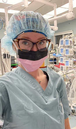 Person in scrubs, surgical cap, glasses, and face mask in an operating room.