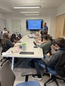 A classroom setting: teacher presents material on a screen to students seated at a large table.