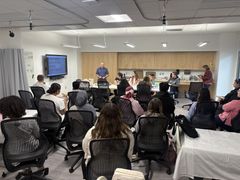 People in a classroom setting, listening to a lecture. A man speaks at the front; students sit in chairs.