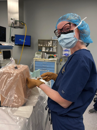 Medical professional in scrubs, mask, glasses, and cap, is working with a medical training dummy in an operating room.