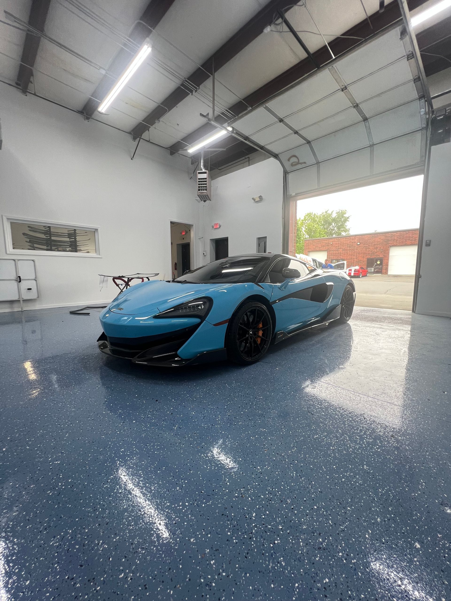 Blue McLaren sports car in a garage with a speckled blue floor, door open to exterior.