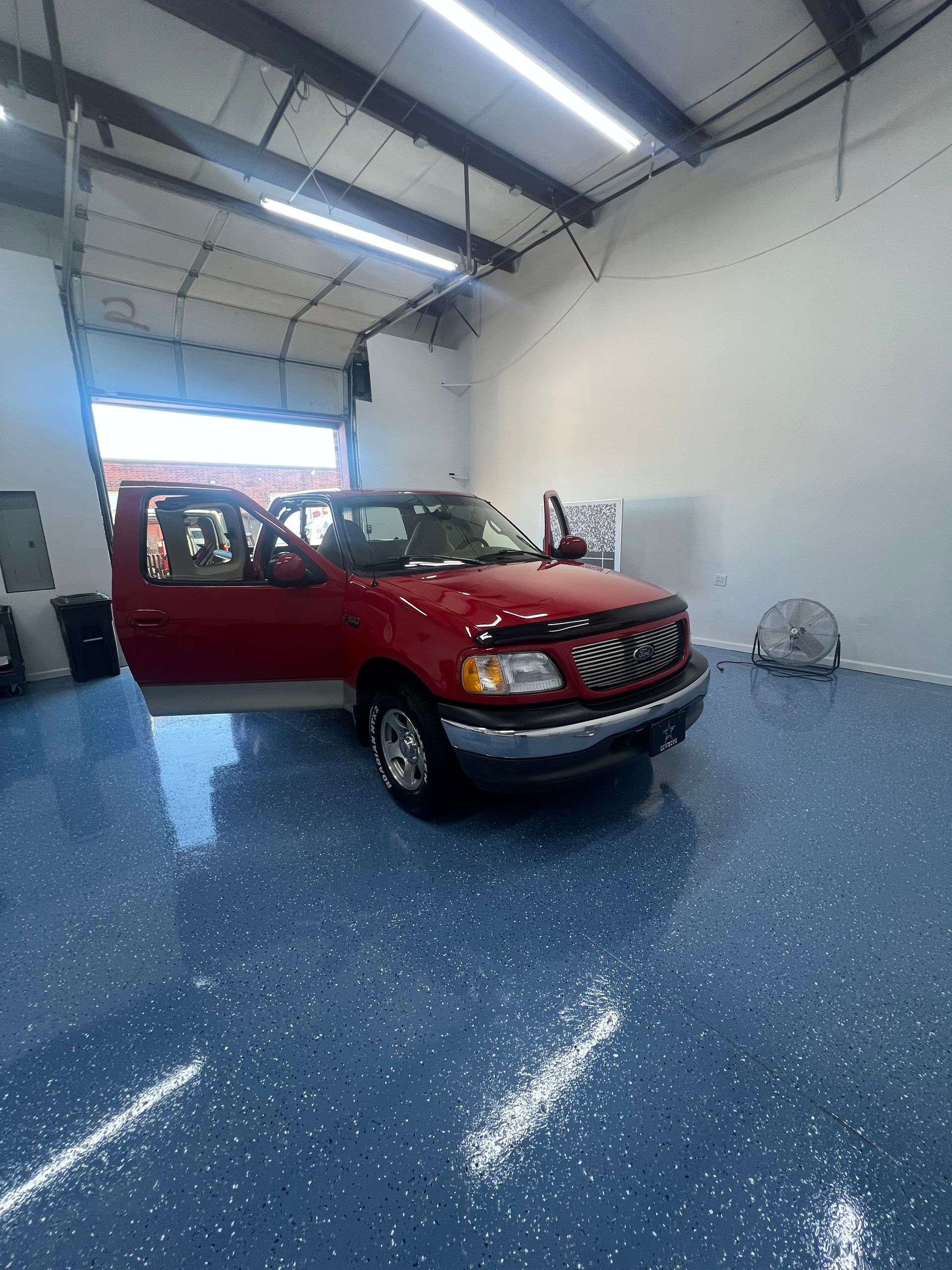 Red Ford truck parked inside a bright shop, door open. Blue speckled floor.