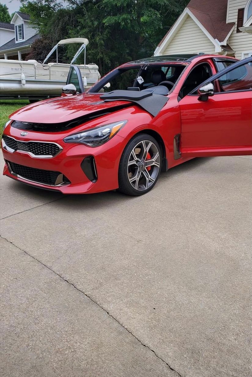 Red Kia Stinger with open doors and damaged hood, parked in front of a boat and house on a driveway.