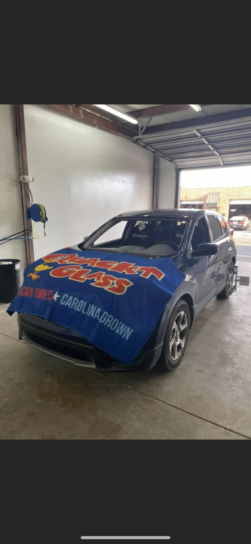 A blue car with a blue cover is in a repair shop. The cover has red and white text.