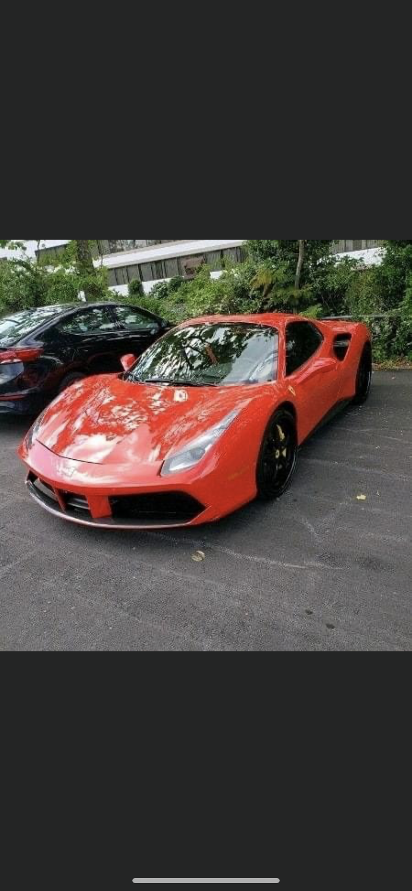Bright orange Ferrari 488 GTB parked on asphalt, black car in background, trees and building visible.