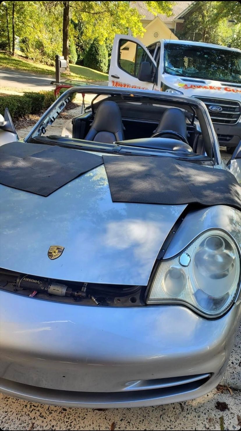 Silver Porsche convertible with damaged front, parked near a white service van.
