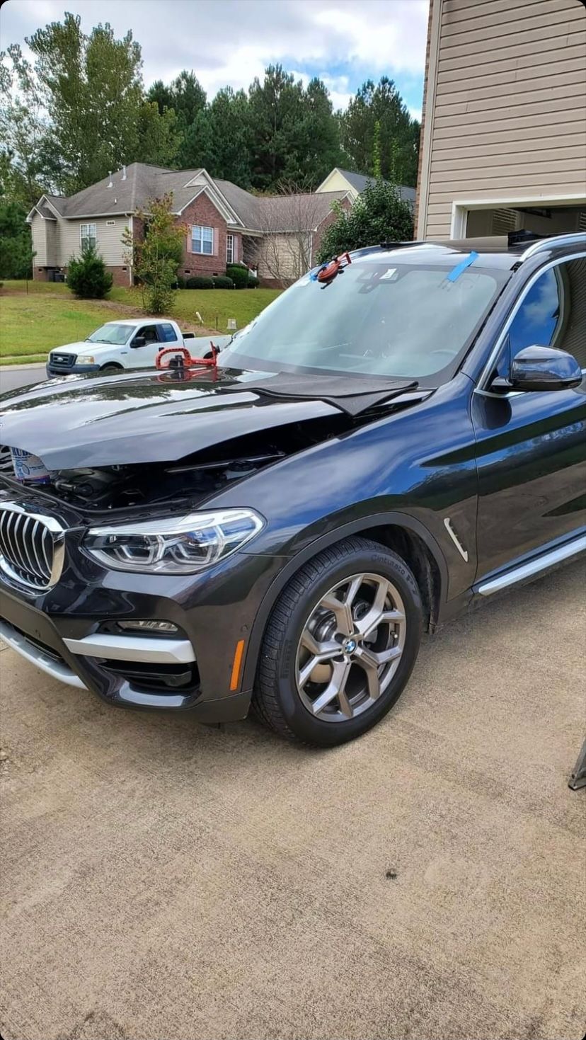 Black BMW SUV with front-end damage parked in a driveway; suburban houses in the background.