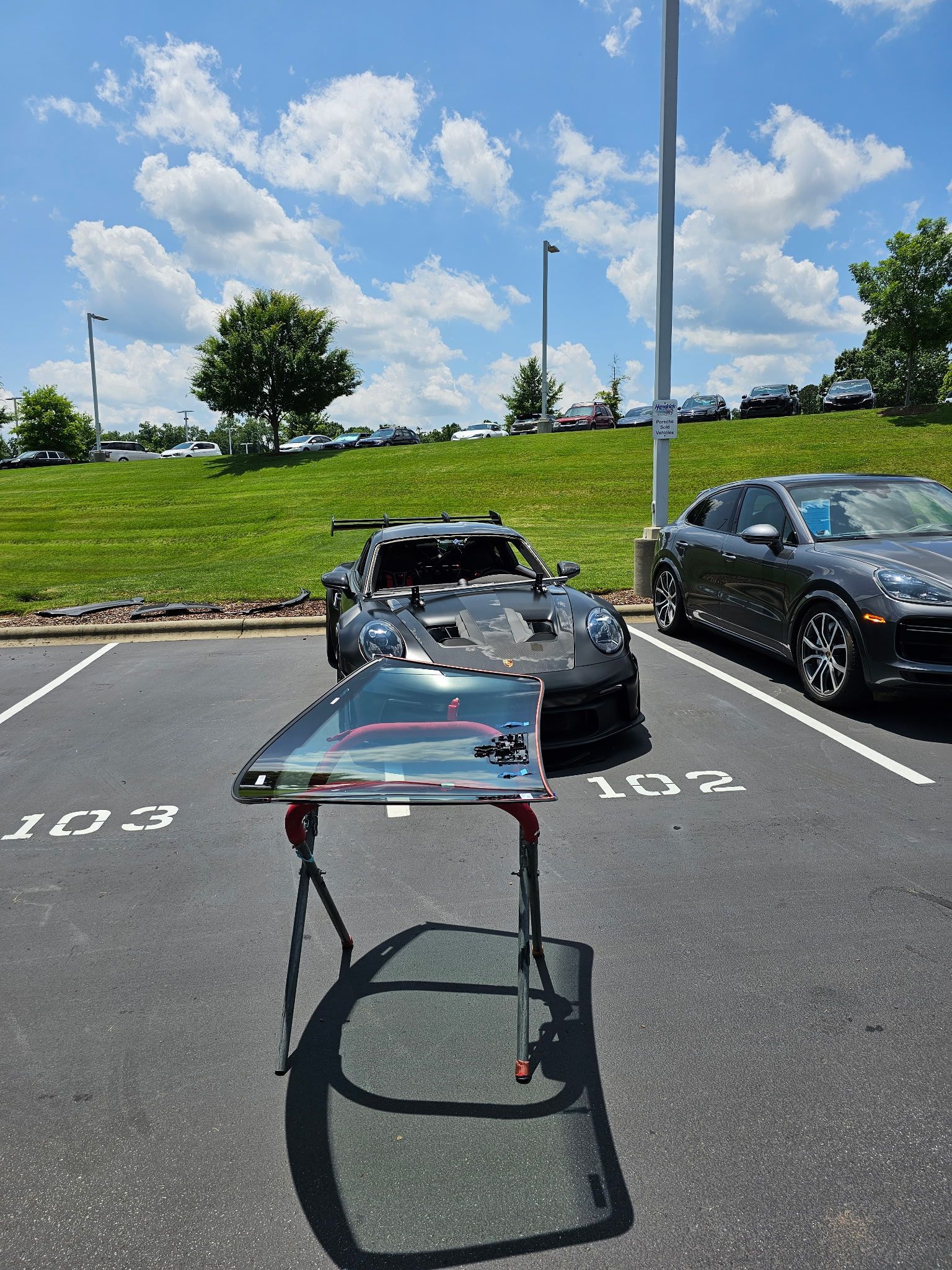A grey sports car and a grey sedan parked in a lot, a windshield rests on a work stand.