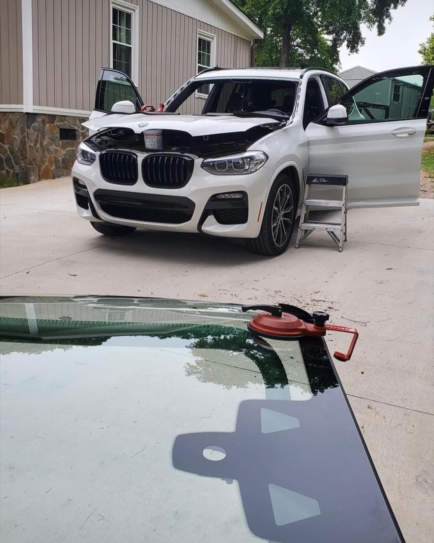 White BMW SUV with open hood and doors, windshield replacement in progress. A new windshield lies on the ground.