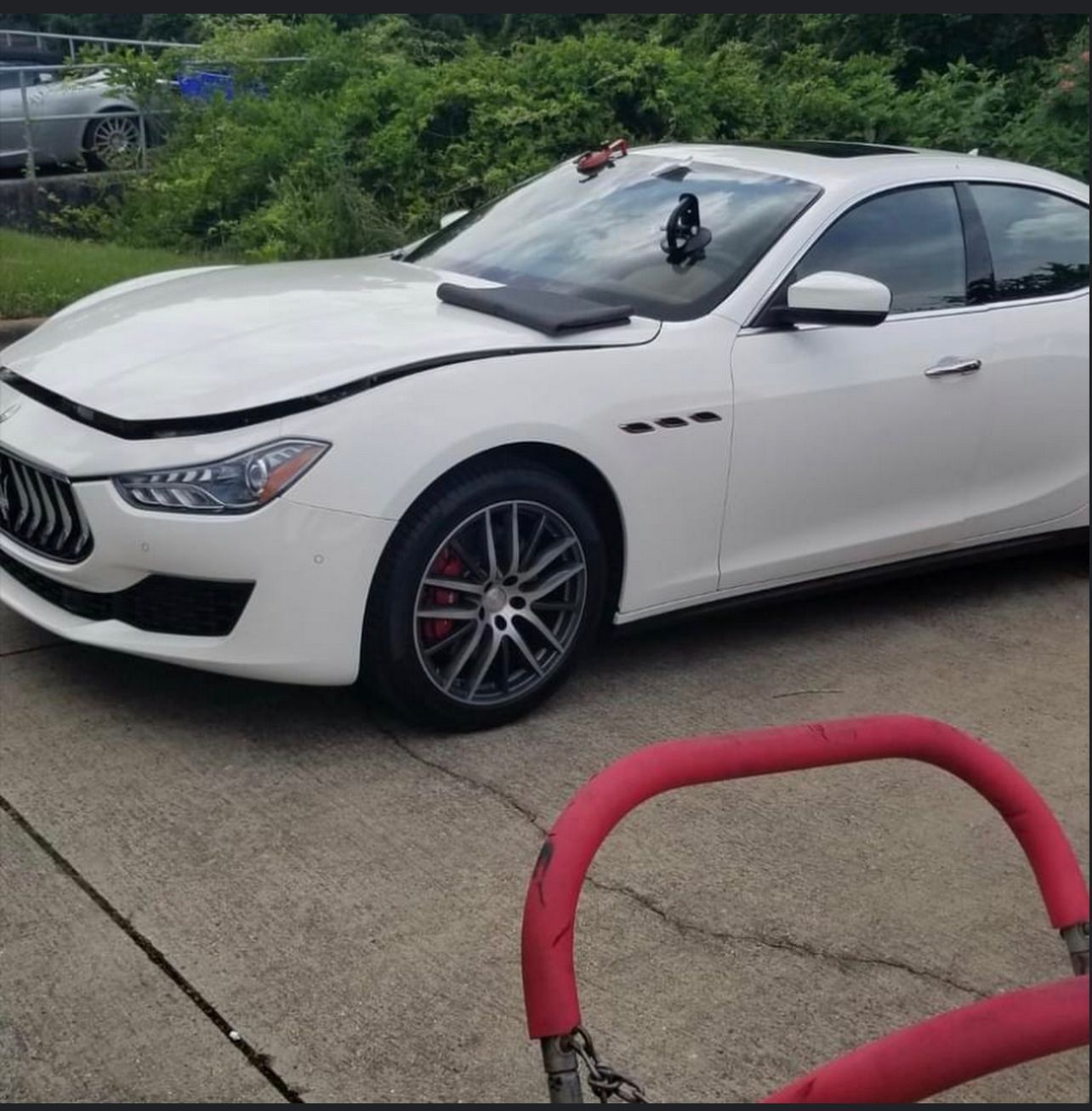 White Maserati sedan with front-end damage parked outside, likely a repair shop, with a red cart in front.