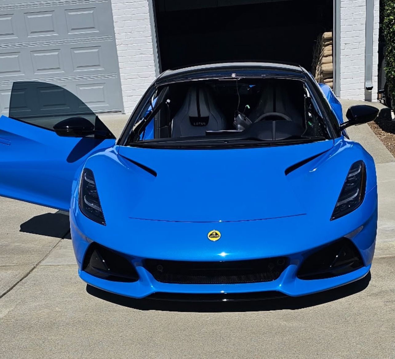 Blue Lotus Emira sports car with the driver's side door open, parked in a driveway.