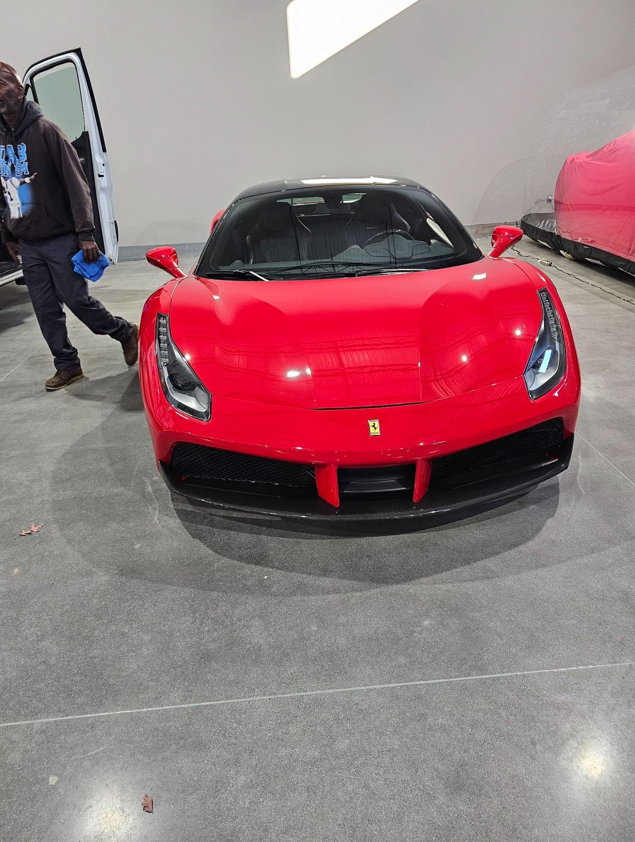 Red Ferrari sports car with black roof, parked in a gray room. Man walks by with open car door.