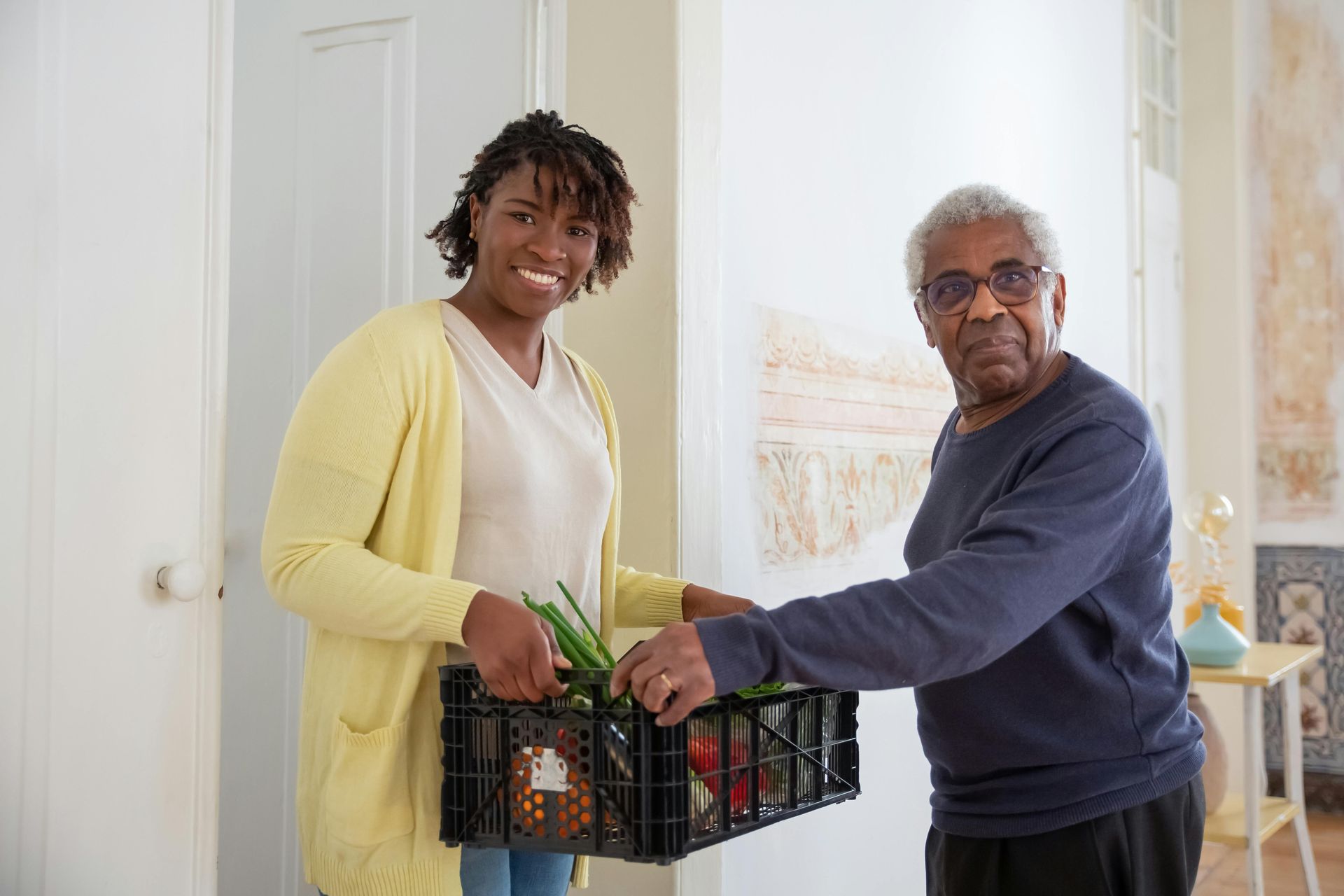 Woman and elderly man smiling, holding a box of groceries in a doorway.