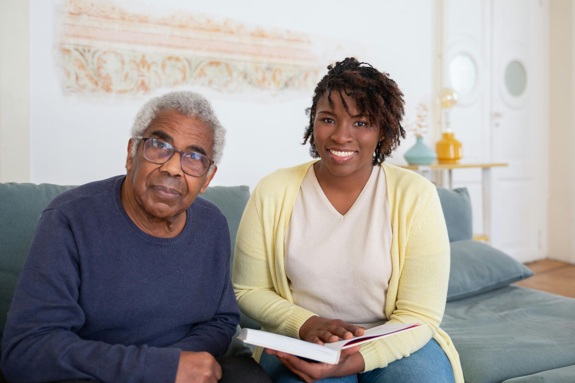 An older Black man and a smiling Black woman sit on a couch. Woman holds a book.