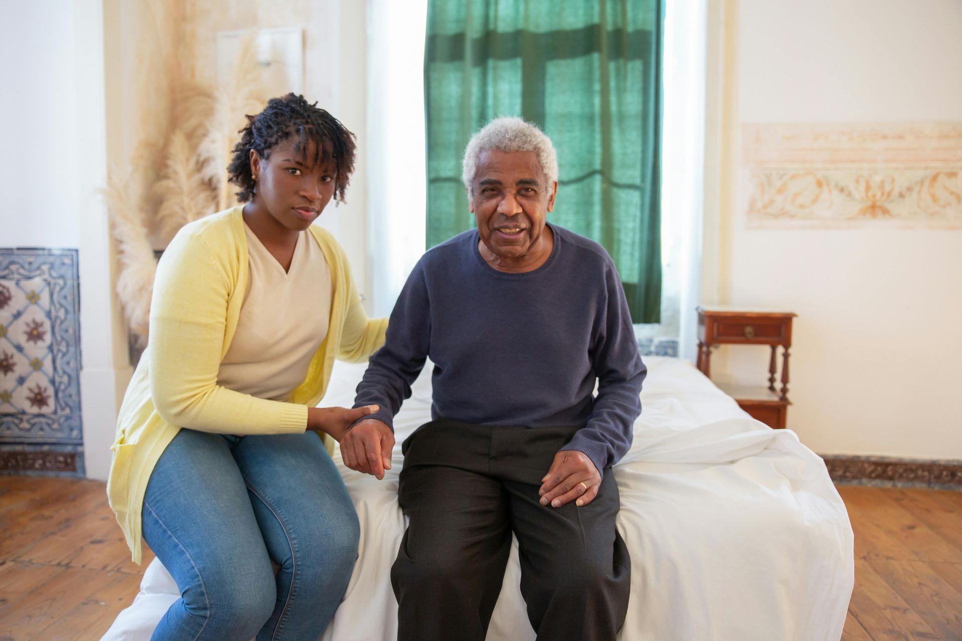 Woman assisting an elderly man to sit on a bed; both look at the camera.