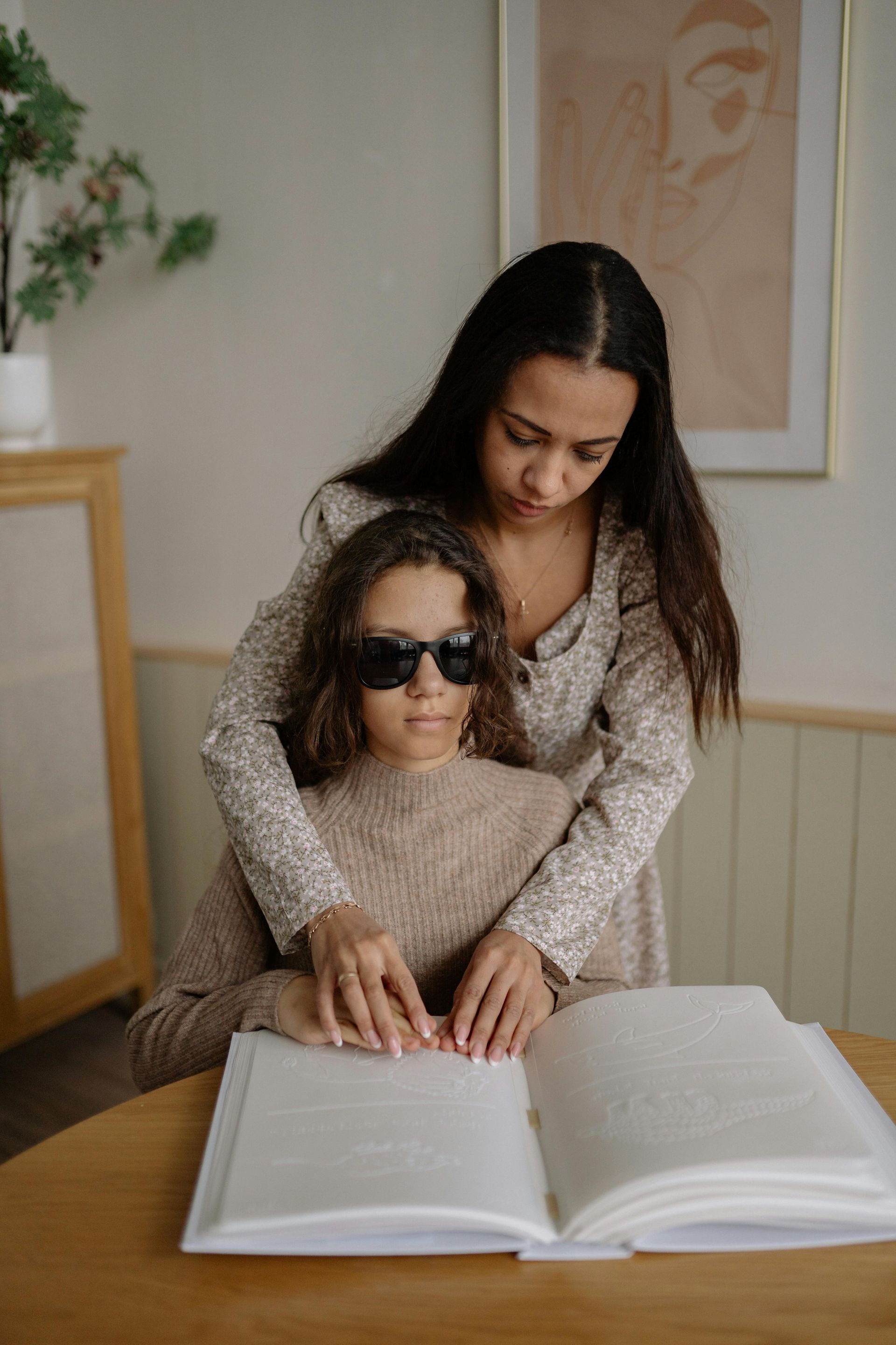 Woman helping another read Braille book at a table.