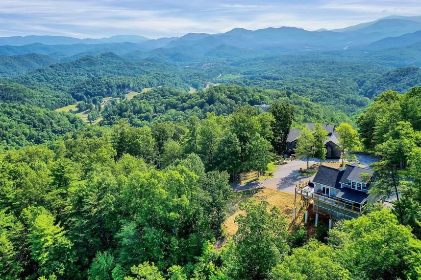 An aerial view of a house in the middle of a forest with mountains in the background.
