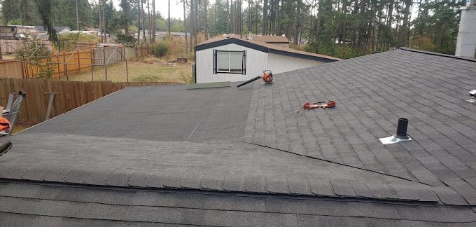 A roof with shingles being installed on it and a house in the background.