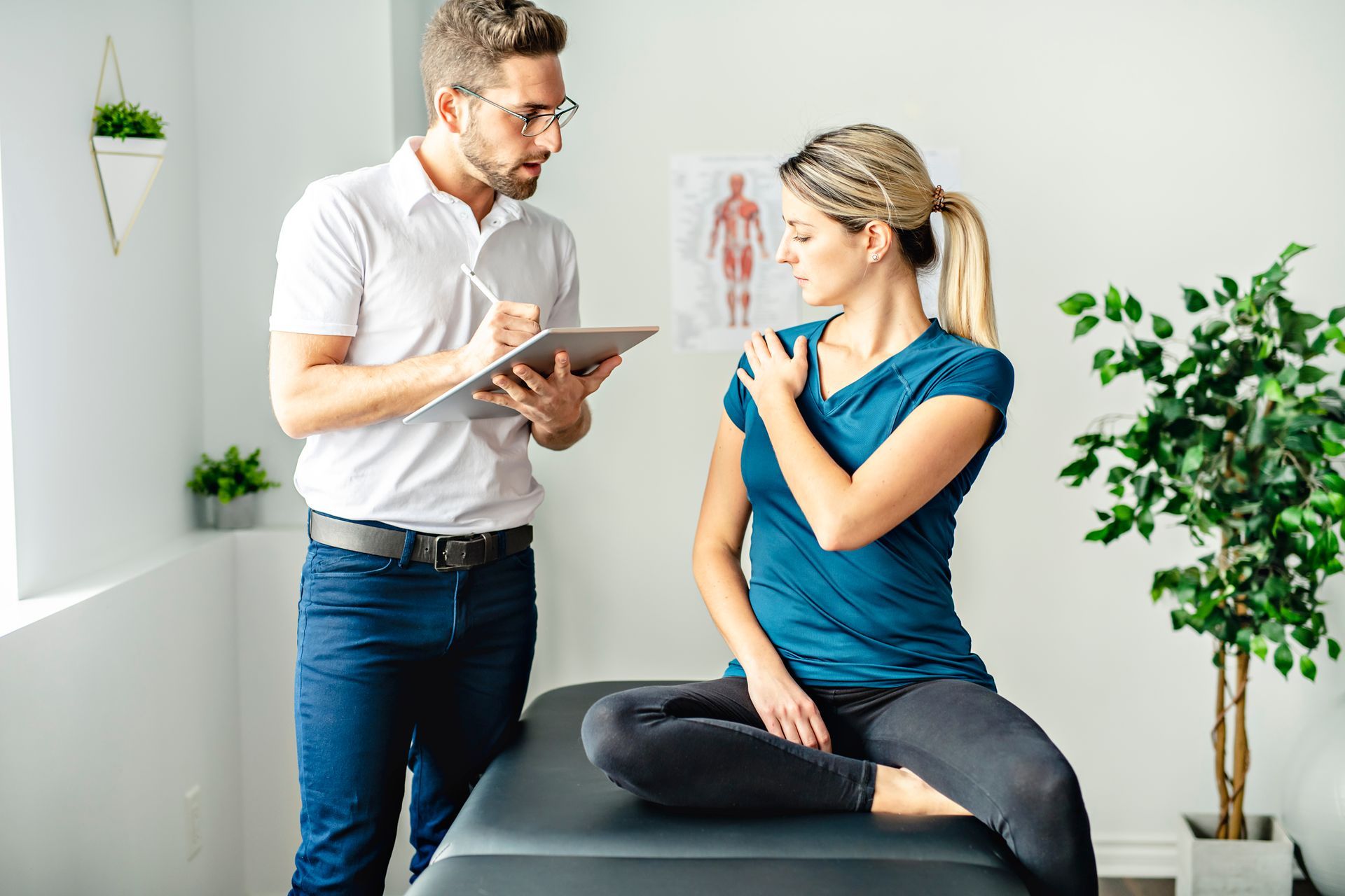 Doctor examining a woman's shoulder, taking notes on a tablet in a bright room.