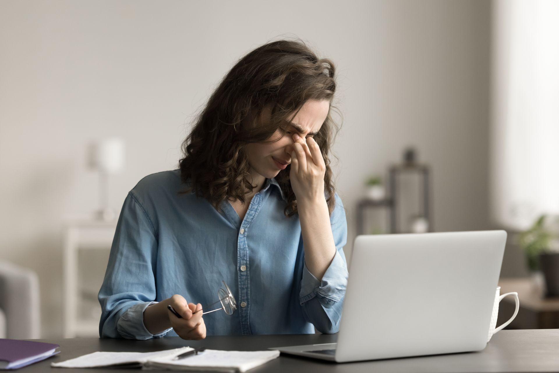 Person at a desk rubbing their eye while working on a laptop in a home office