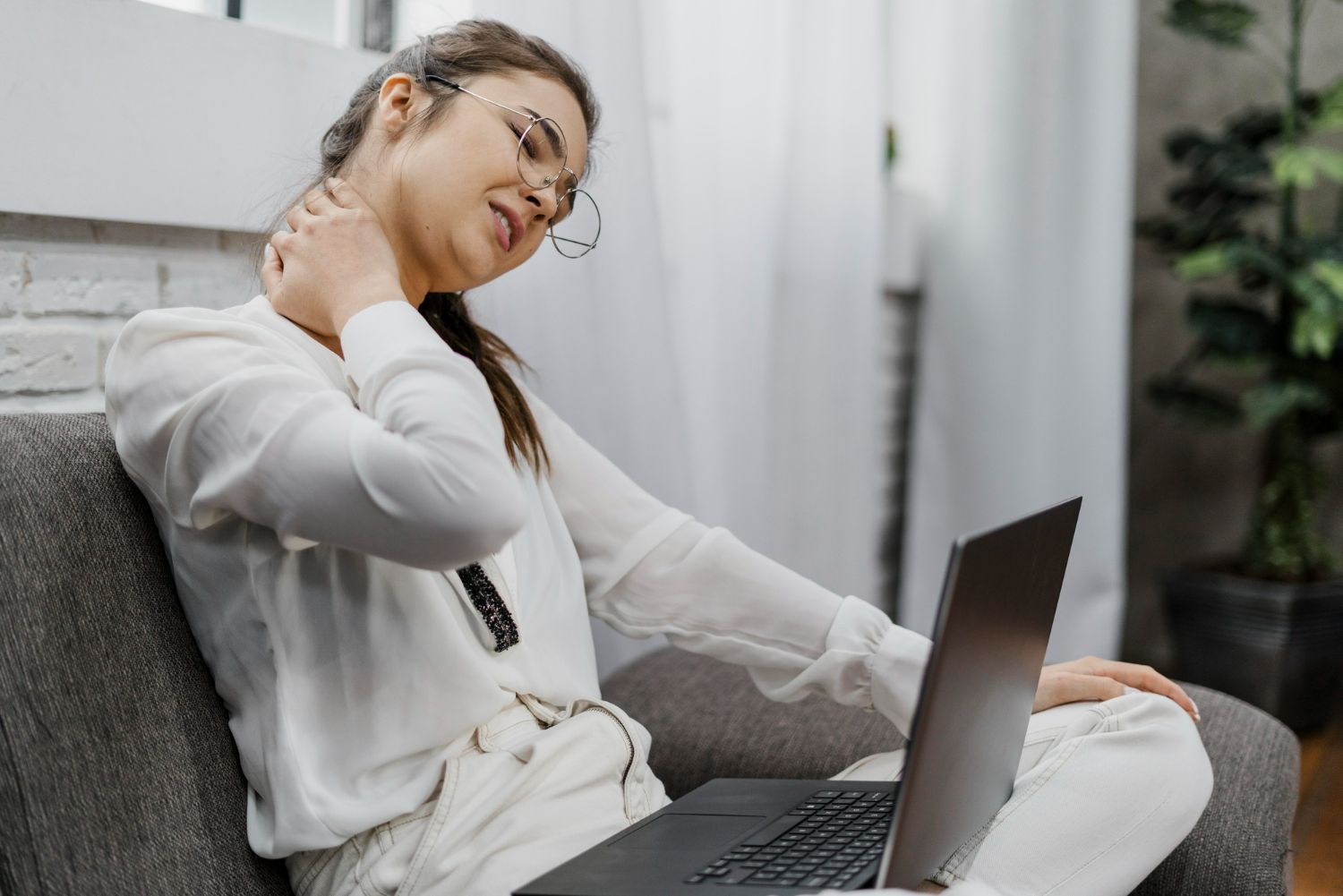 A woman is sitting in a chair with a laptop and holding her neck.