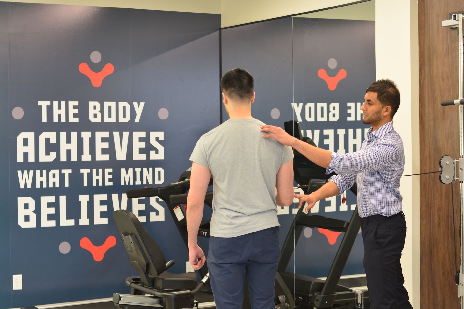 Two men on a treadmill in a gym, with a wall slogan reading “The body achieves what the mind believes.”
