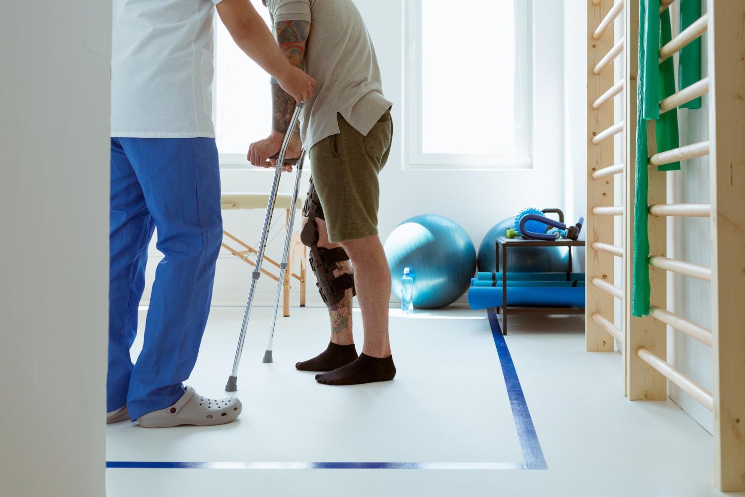 A nurse is helping a man with crutches walk in a gym.