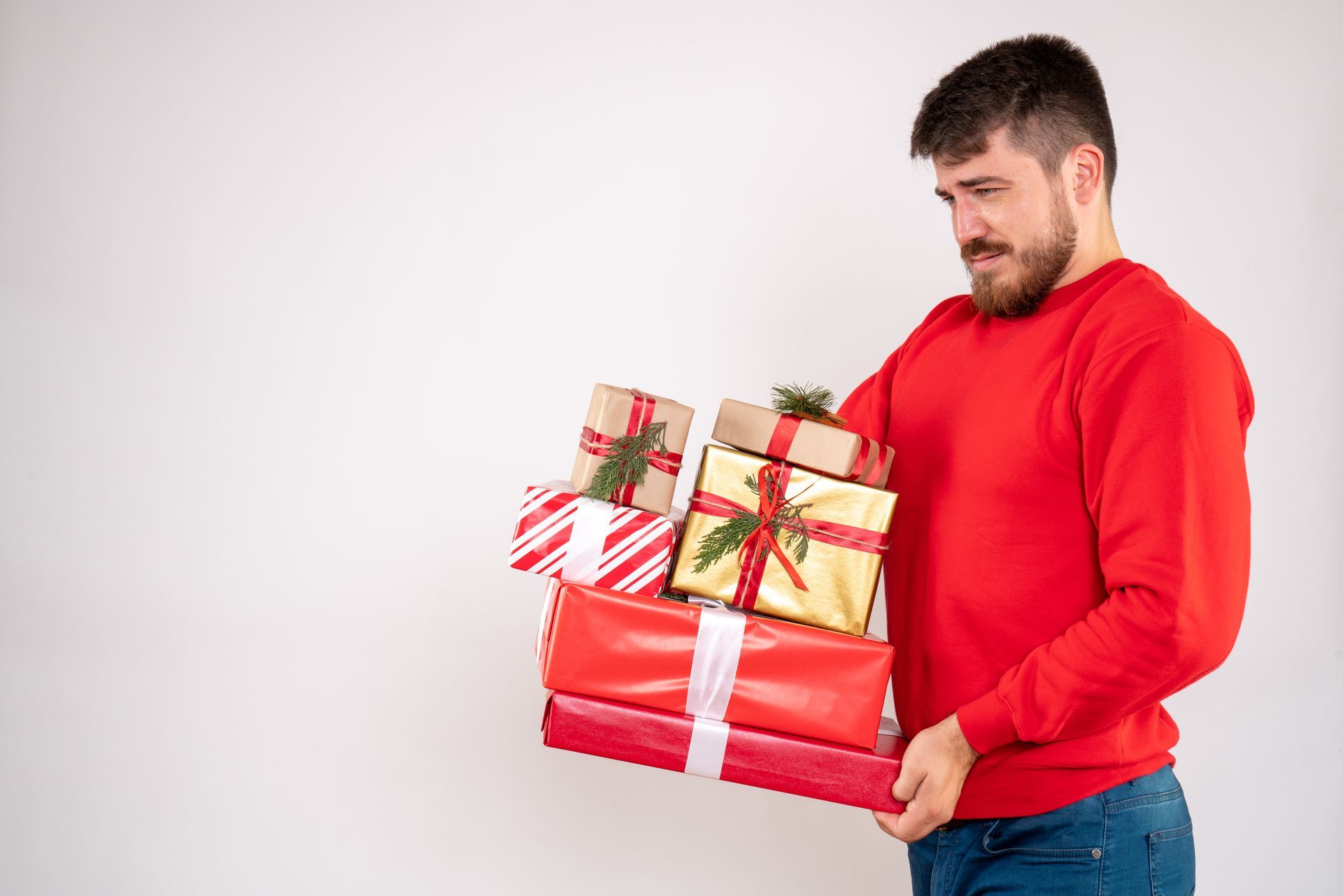 Man in red sweater struggling to hold a stack of wrapped Christmas gifts, white background.
