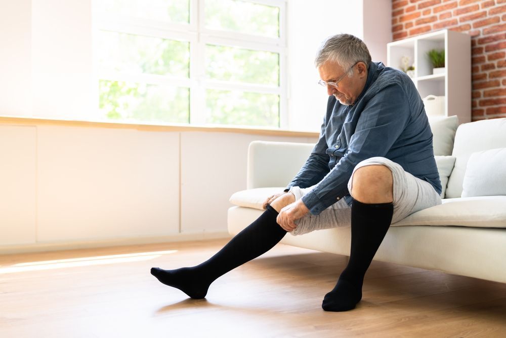 An older man is sitting on a couch putting on compression socks.