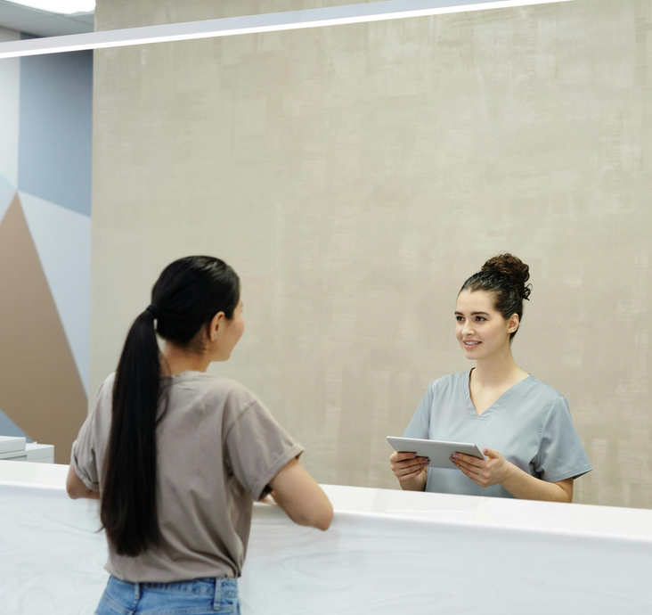 Woman speaking with a receptionist at a white reception desk, in a modern, light-filled office.