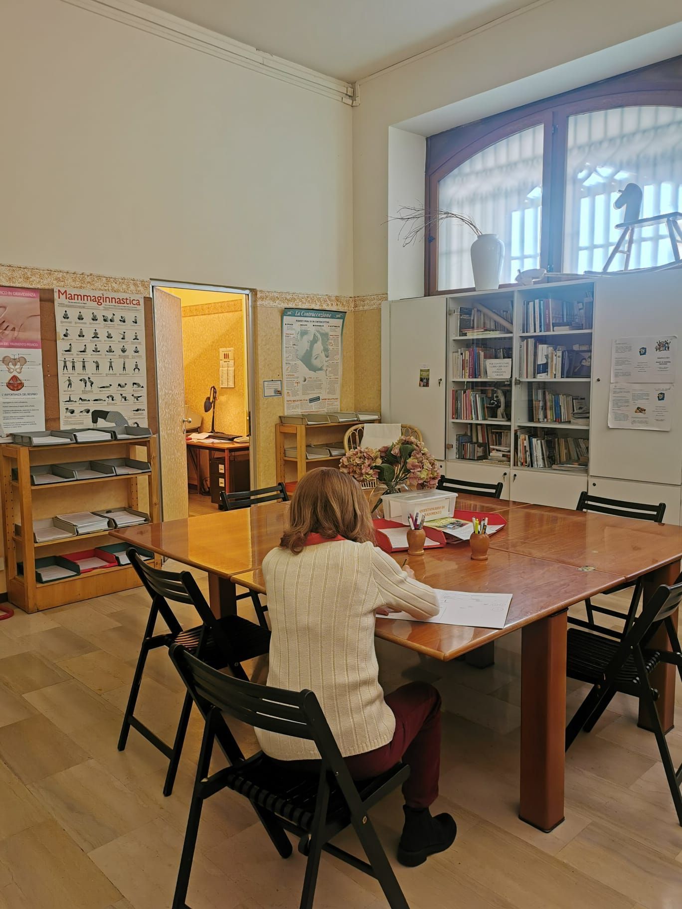 Donna seduta a un tavolo che scrive in una stanza con una libreria, una porta aperta e una finestra.
