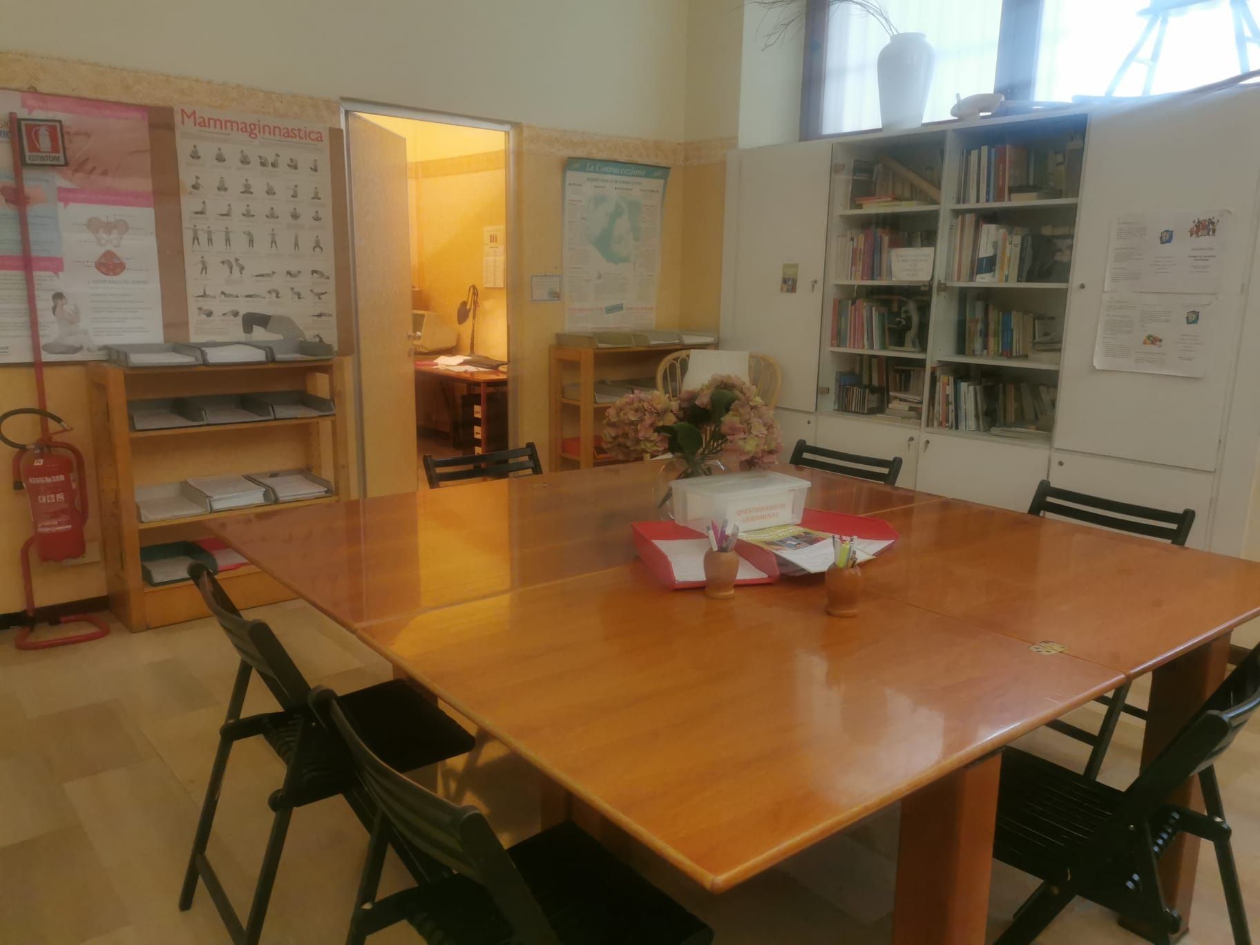 Stanza con grande tavolo, sedie, librerie e porta che conduce a un'altra stanza.