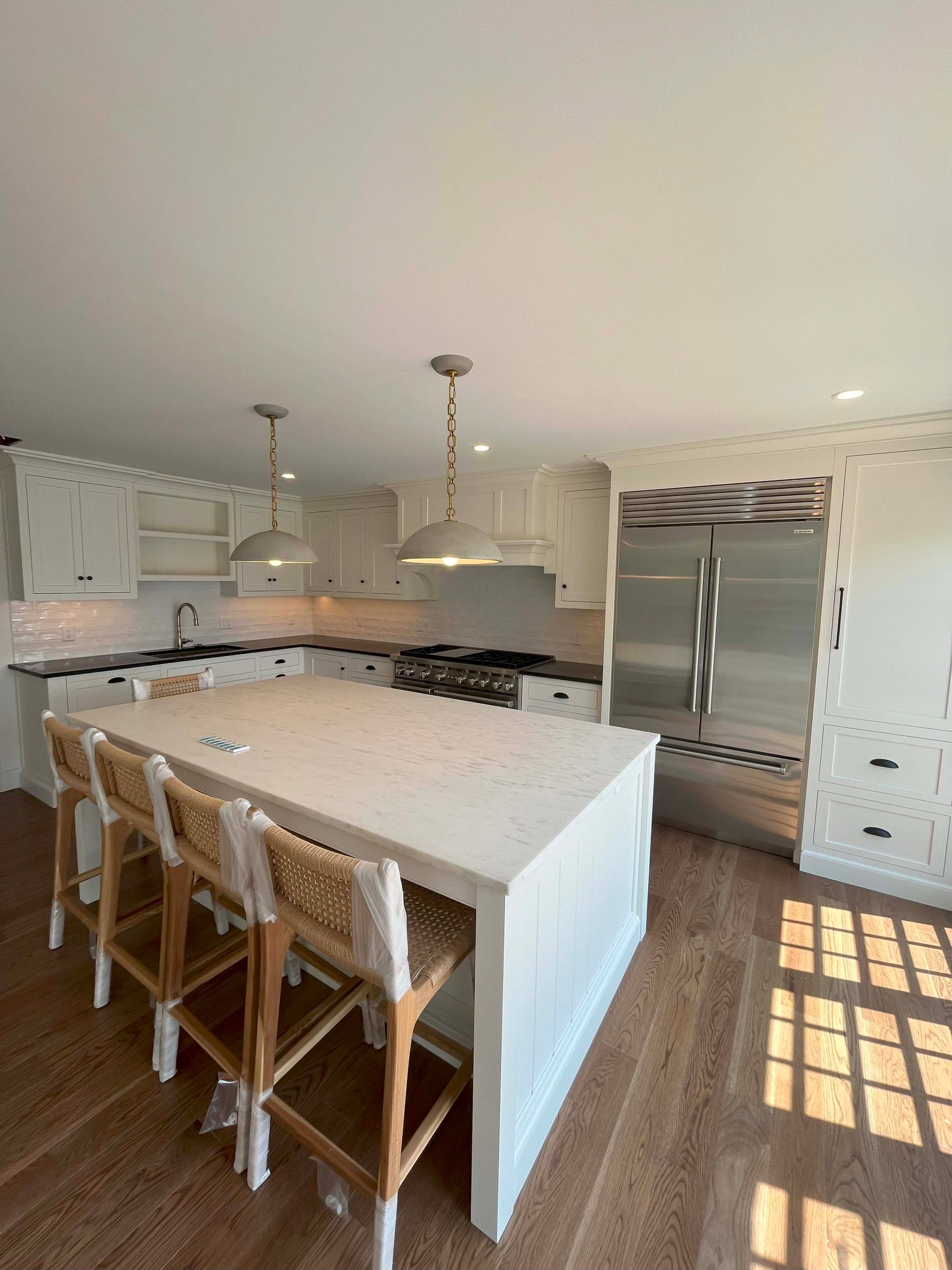 White kitchen with island and stools; stainless steel fridge; pendant lights.