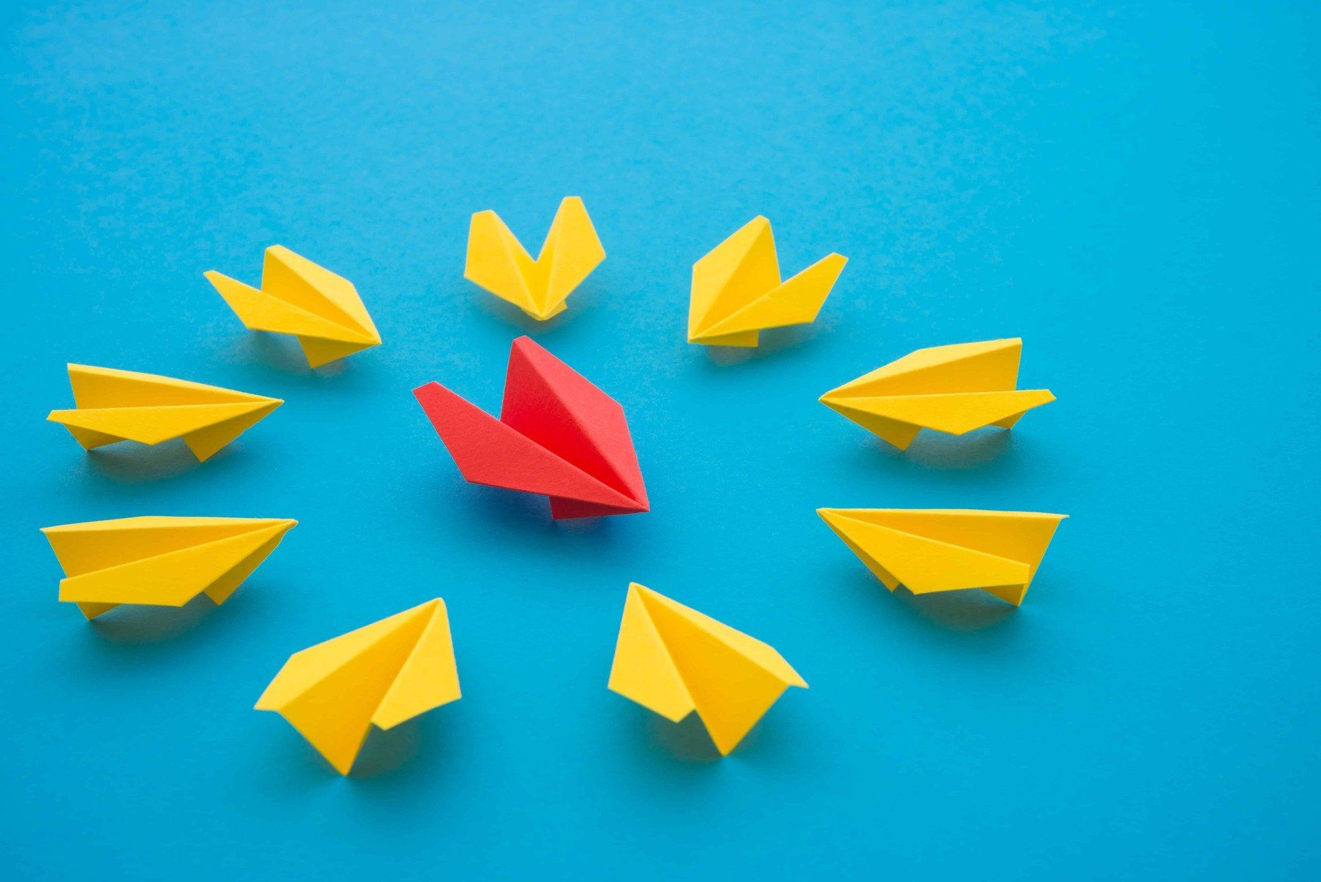 A red paper airplane is surrounded by yellow paper airplanes on a blue background.