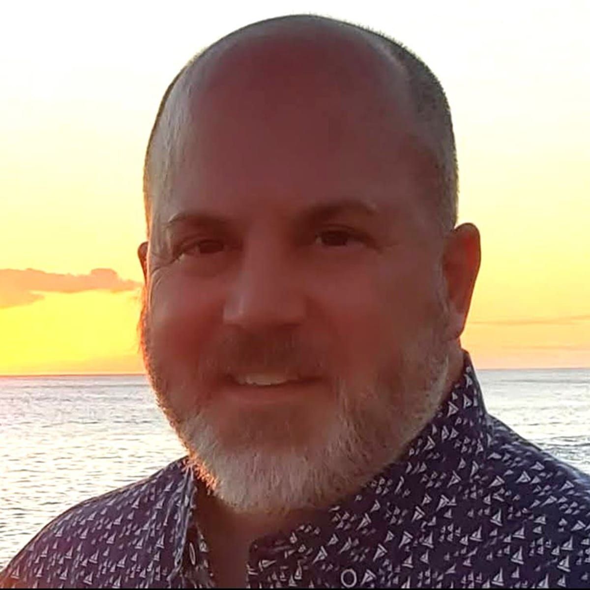 A man with a beard is smiling in front of the ocean at sunset.