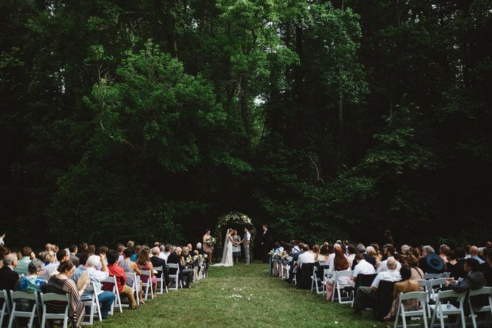 A large group of people are sitting in white chairs at a wedding ceremony in the woods.
