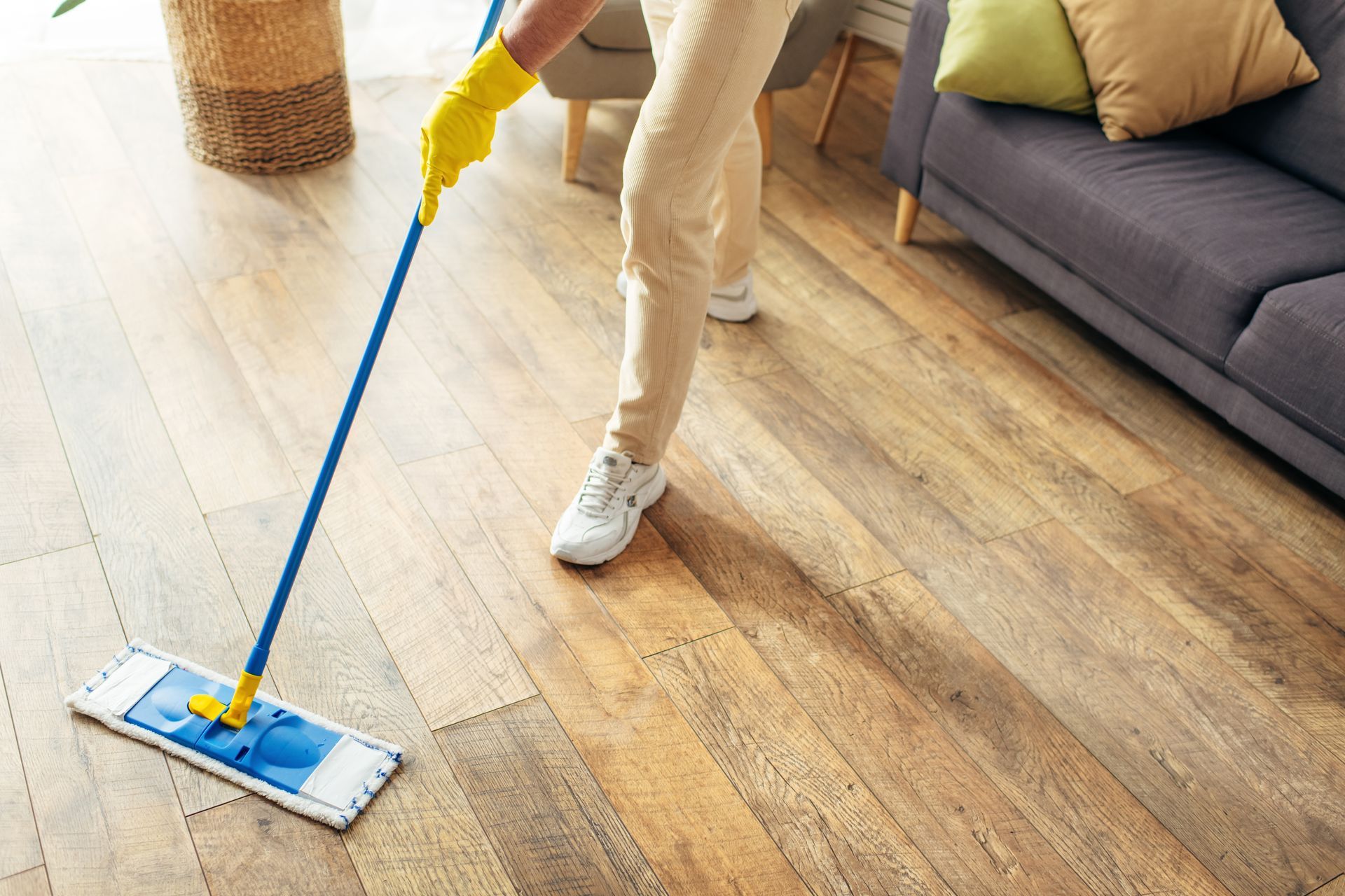 Person mopping a wooden floor in a living room, wearing yellow gloves and white sneakers.