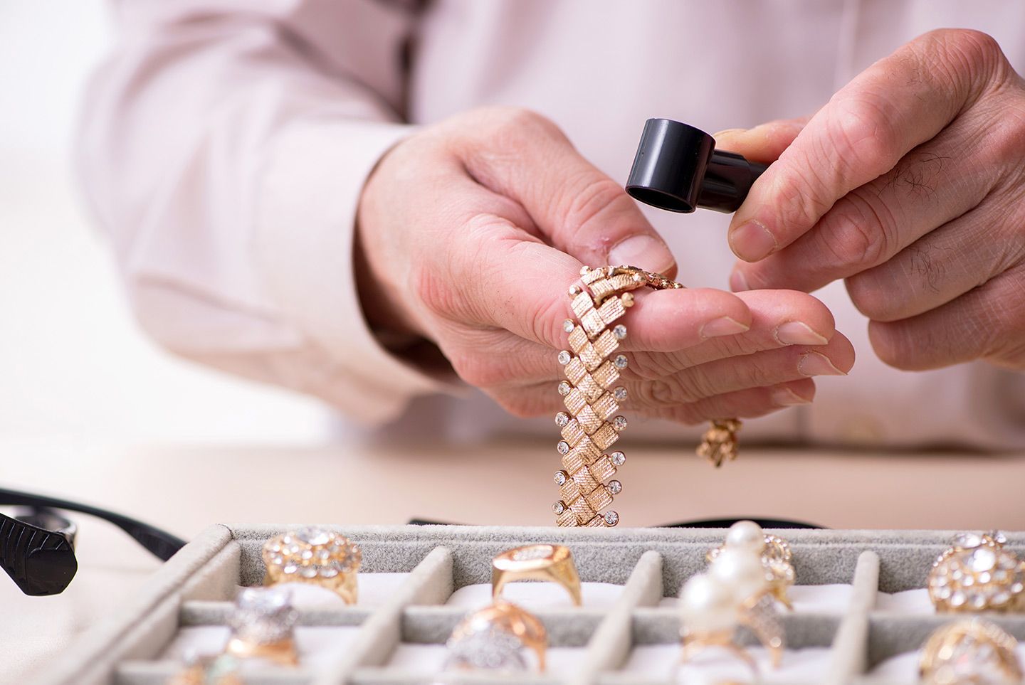 Jeweler examines gold bracelet with a magnifying glass, jewelry tray in foreground.