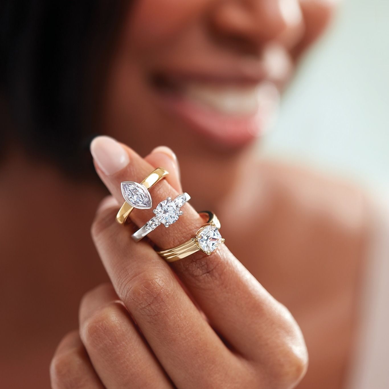 Woman's hand holding three diamond rings: pear, round, and heart-shaped. Smiling with a blurred background.