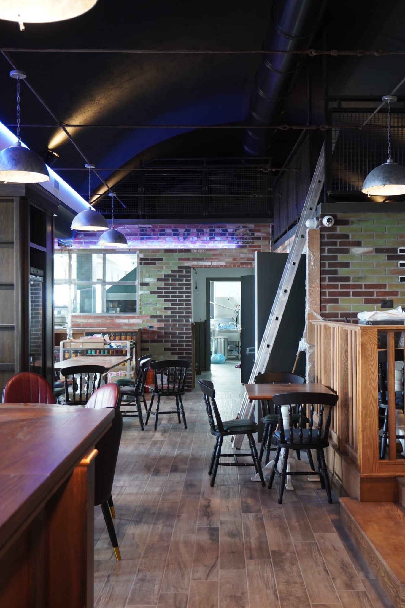 Interno di un ristorante con tavoli in legno, sedie nere, muro in mattoni e illuminazione dall'alto.