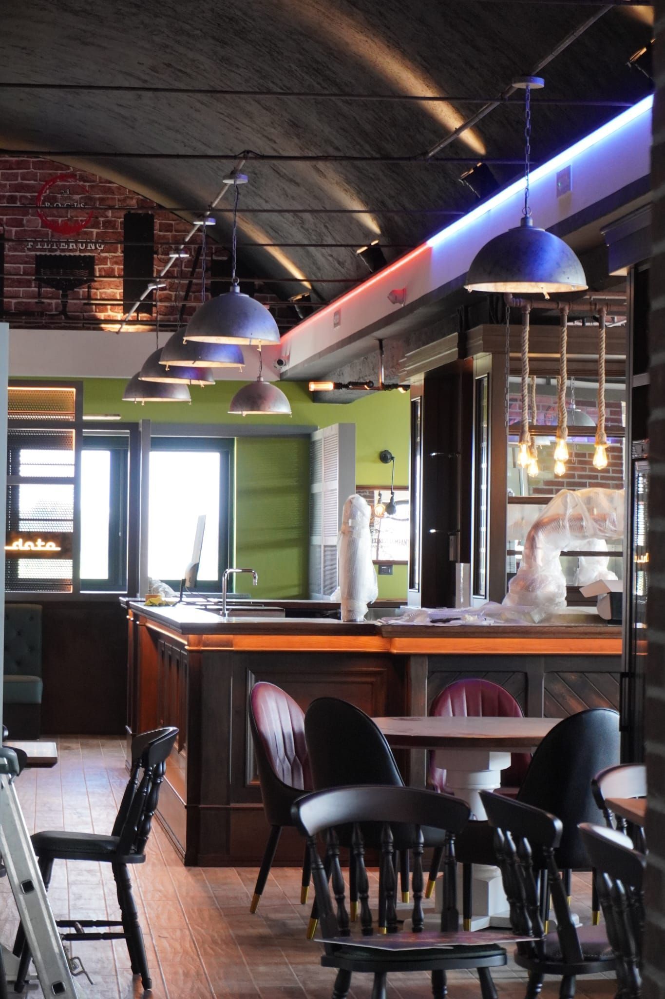Interno di un pub con bancone, tavoli, sedie e luci sospese. Sono presenti anche pareti in mattoni e una parete verde brillante.