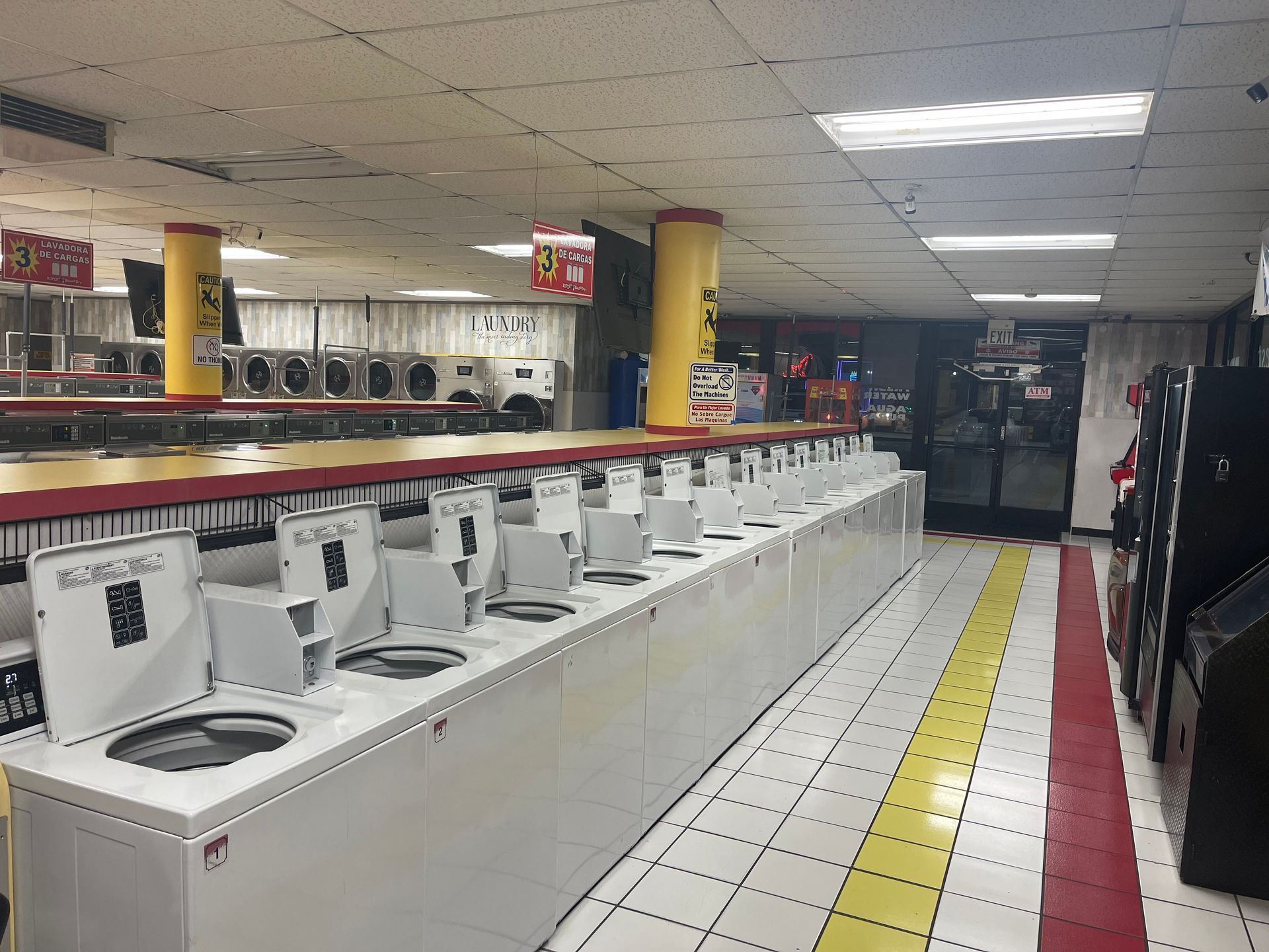 A laundromat interior with rows of washing machines. White appliances, yellow pillars, and a red and yellow floor.